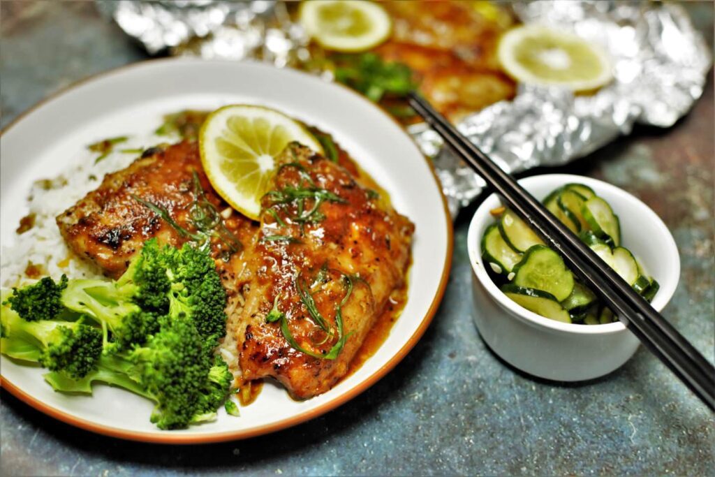 Close-up photo of a piece of salmon with a golden-brown glaze made of miso and maple syrup laying on rice. The salmon is placed on a white plate with some escallion garnish sprinkled on top and a few pegs of broccoli and cucumber on the side.